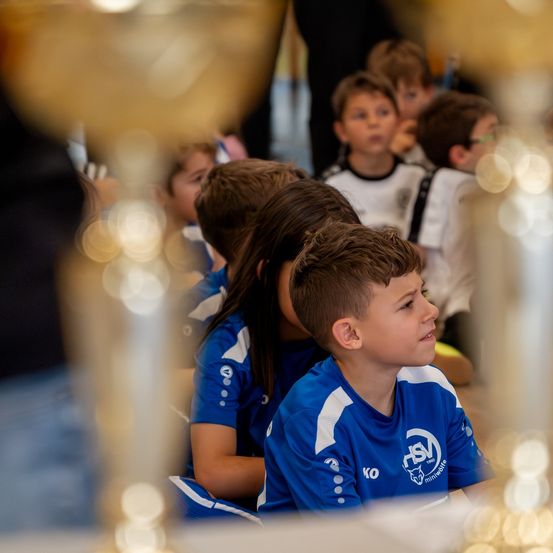 Mehrere Kinder sitzen in einer Reihe, tragen blaue und weiße Trikots, mit einer verschwommenen Ansicht von Pokalen und einer weiteren Person im Hintergrund.