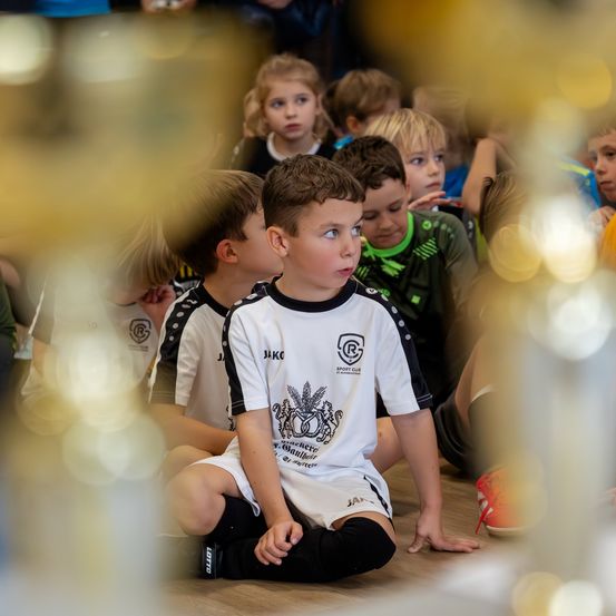 Eine Gruppe von Kindern in Sportuniformen sitzt auf dem Boden und schaut interessiert etwas an. Der Junge in der Mitte trägt ein weißes Shirt mit Logo.