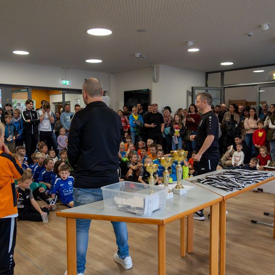 Zwei Männer stehen hinter einem Tisch mit Trophäen, während eine Menge Kinder und Erwachsene zusieht. Der Raum hat Holzböden und weiße Wände mit Lampen an der Decke.