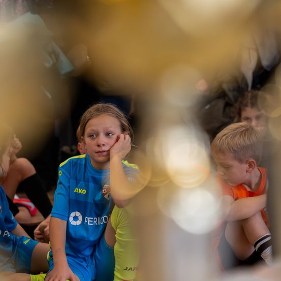 Eine Gruppe von Kindern sitzt auf dem Boden, ein Mädchen in Blau und Orange, ein anderes in Orange, beide schauen nach unten, umgeben von verschwommenen Figuren.