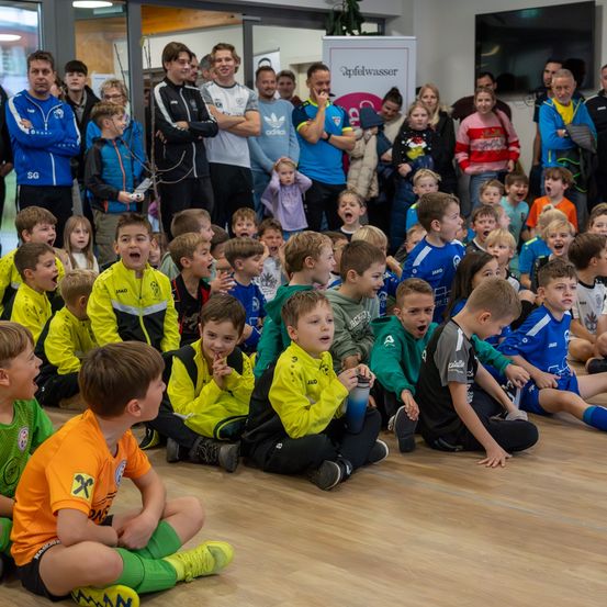 Junge Fußballspieler in gelben, grünen und blauen Trikots sitzen auf dem Boden, während ein Mann in einem blauen Jacke ein Schild hält. Andere Menschen, einschließlich Erwachsene, stehen hinter ihnen, und ein Fernseher ist an der Wand montiert.