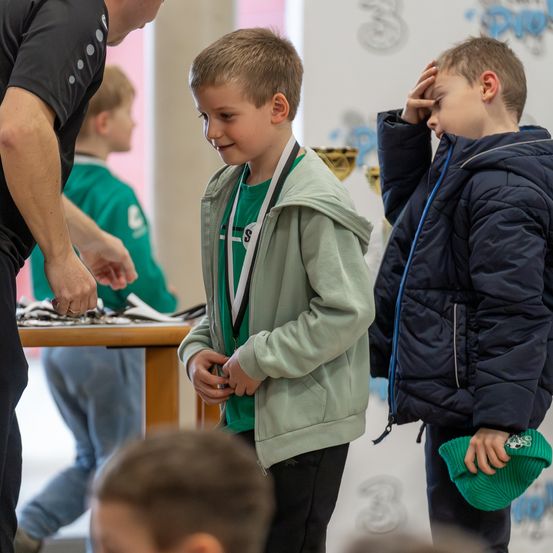 Ein junger Junge mit einer Medaille um den Hals steht neben einem anderen Jungen mit der Hand auf der Stirn. Ein Mann verteilt Gegenstände von einem Tisch.