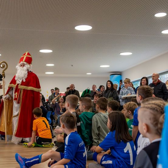 Eine Gruppe von Kindern und Erwachsenen versammelt sich in einem Raum. Ein Mann, der als Weihnachtsmann verkleidet ist, steht vor ihnen. Die Kinder sitzen auf Stühlen und schauen ihn an. Einige von ihnen machen Fotos mit ihren Handys. Der Raum hat einen Holzboden und eine weiße Decke.