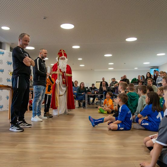 Eine Gruppe von Kindern in blauen Uniformen sitzt auf dem Boden, wahrscheinlich in einer Turnhalle. Sie beobachten einen älteren Mann, der als Weihnachtsmann gekleidet ist. Zwei Männer in Schwarz stehen vor ihnen. Dahinter sitzt eine Menschenmenge auf Stühlen, einige stehen. An der Decke hängen Lampen.