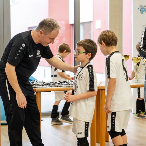 Ein Mann in einem schwarzen Shirt spricht mit zwei Jungen in weißen und schwarzen Trikots. Sie stehen vor einem Tisch mit Trophäen und im Hintergrund befindet sich eine Glaswand.
