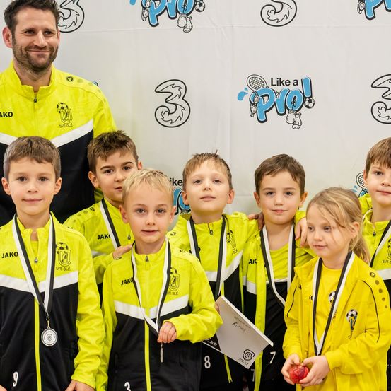 Ein lächelnder Mann steht hinter einer Gruppe von Kindern in gelben und schwarzen Trikots mit Medaillen und posiert für ein Foto. Die Kinder sehen aufgeregt aus und einige halten kleine Gegenstände. Der Hintergrund zeigt Logos und Text.