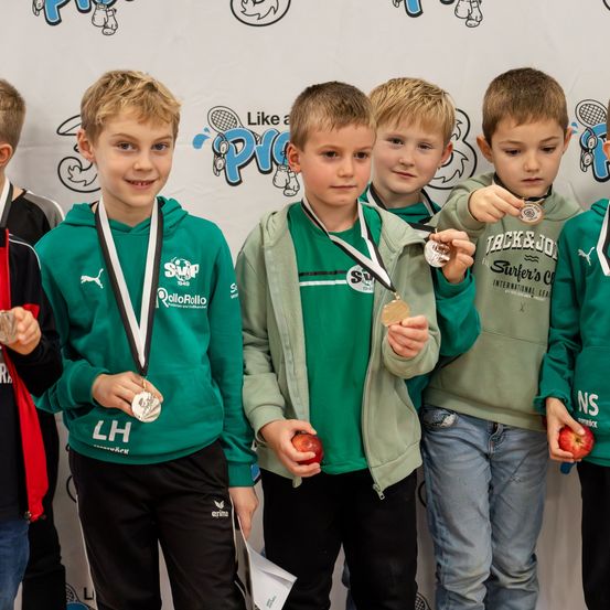 Fünf Jungen in Sporttrikots halten Äpfel und Medaillen und stehen in einer Reihe, mit einem weißen Hintergrund und Logos dahinter.
