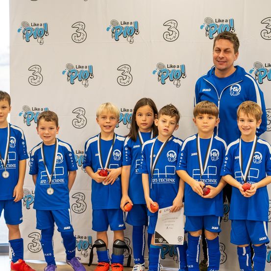 Eine Gruppe von Kindern in blauen Uniformen mit Medaillen posiert für ein Foto mit einem Trainer. Der Hintergrund zeigt den Text 'Wie ein Profi!' und die Zahlen 3.