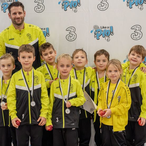 Eine Gruppe junger Athleten in gelben und schwarzen Uniformen mit Medaillen, die vor einem Hintergrund mit Logos und Nummern stehen. Sie posieren für ein Foto mit einem Mann in einem gelben Jackett.