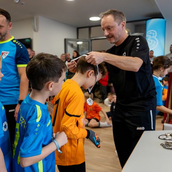 Ein Mann legt einem Jungen eine Medaille um den Hals. Andere Kinder und Erwachsene schauen zu. Der Raum hat einen Holzboden und weiße Wände.