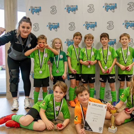 Eine Gruppe junger Fußballer in grünen Trikots steht mit einem Trainer, der Pokale und Äpfel hält. Sie posieren für ein Foto vor einer Kulisse mit Sponsorenlogos.
