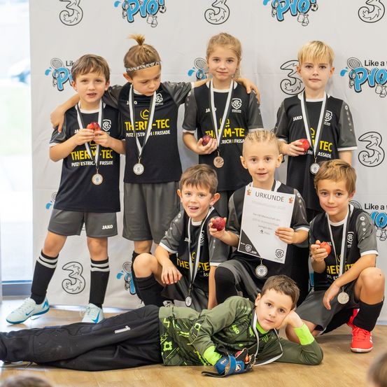 Eine Gruppe junger Athleten, die Sporttrikots und Medaillen tragen, posiert für ein Foto. Sie halten Äpfel und stehen vor einem weißen Hintergrund mit einem Logo.