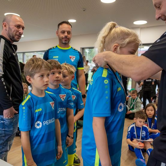 Erwachsene und Kinder in blauen Uniformen versammeln sich in einem Raum. Ein Mann hilft einem Mädchen mit einer Medaille. Andere Kinder sitzen im Hintergrund auf dem Boden.