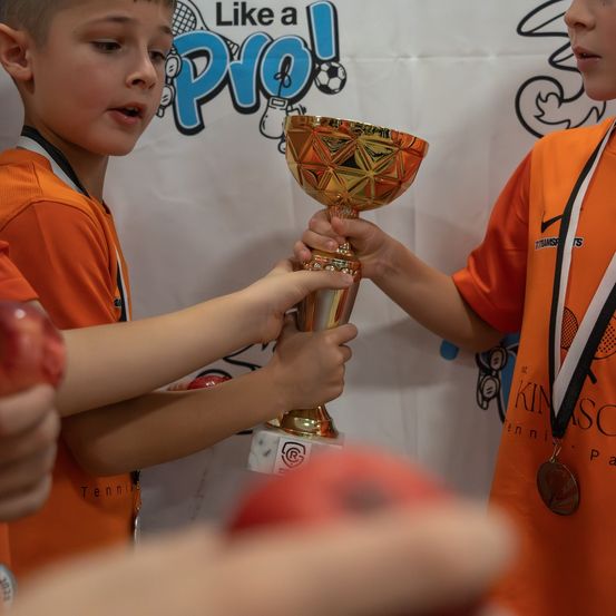 Zwei junge Jungen in orangefarbenen Trikots halten einen Pokal. Sie sehen aufgeregt und glücklich aus. Hinter ihnen ist ein Hintergrund mit Text und Bildern.