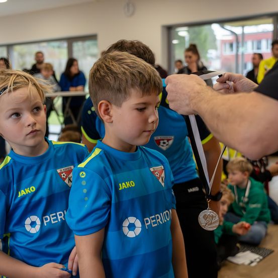 Zwei junge Jungen in blauen Fußballtrikots mit dem Wort 'Peri' darauf erhalten eine Silbermedaille von einem Erwachsenen in einem Raum mit mehreren Personen.