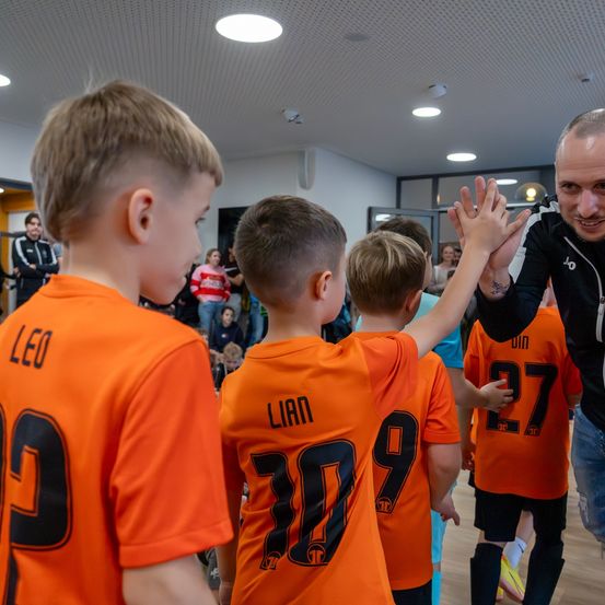 Ein lächelnder Mann in einem schwarzen Jackett schüttelt die Hände mit einer Gruppe von Jungen in orangefarbenen Trikots mit Nummern. Die Jungen stehen in einem Raum mit anderen Leuten im Hintergrund.