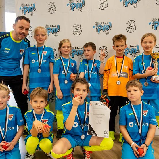 Eine Gruppe von Kindern in Sportuniformen, einige mit Medaillen und Trophäen, posieren für ein Foto. Sie stehen vor einem weißen Hintergrund mit Sportlogos.