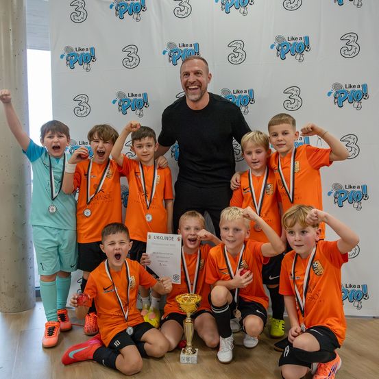 Eine Gruppe junger Jungen in orangefarbenen Trikots feiert mit einem Pokal und Medaillen und posiert mit einem Mann in einem schwarzen T-Shirt vor einem dekorierten Hintergrund.