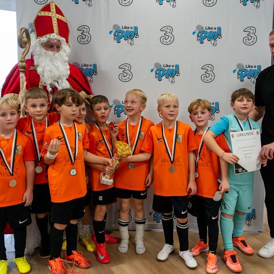 Eine Gruppe junger Fußballer in orangefarbenen Trikots steht mit Medaillen und Trophäen, hinter ihnen ein Mann als Weihnachtsmann verkleidet.