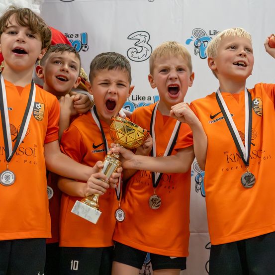 Eine Gruppe junger Jungen in orangefarbenen Trikots und Medaillen feiert ihren Sieg. Sie stehen zusammen, halten eine goldene Trophäe und zeigen stolz ihre Medaillen.