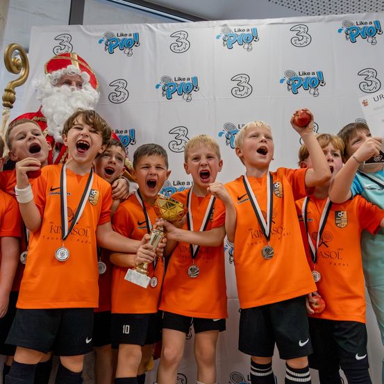 Eine Gruppe junger Jungen in orangefarbenen Trikots mit Medaillen um den Hals posiert für ein Foto mit einem Pokal. Sie tragen alle passende Uniformen und scheinen einen Sieg zu feiern. Hinter ihnen steht ein Mann in Santa-Claus-Kleidung, der einen Apfel hält und lächelt.