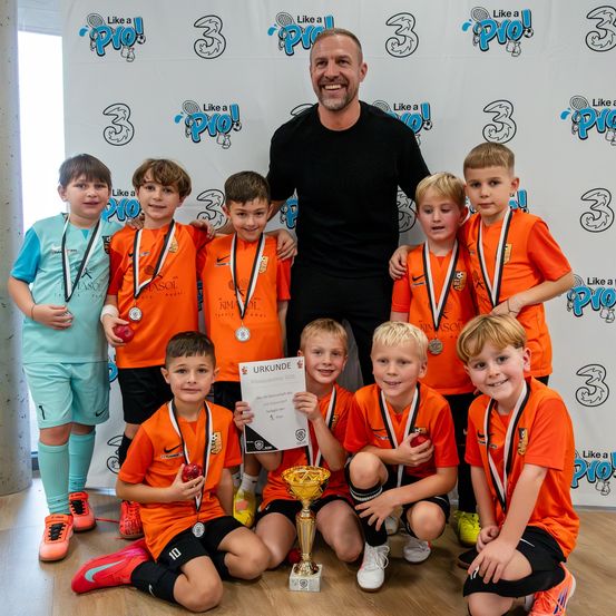 Eine Gruppe junger Fußballer in orangefarbenen Trikots posiert mit ihrem Trainer, hält Medaillen und einen Pokal und steht vor einem weißen Hintergrund mit Logos.
