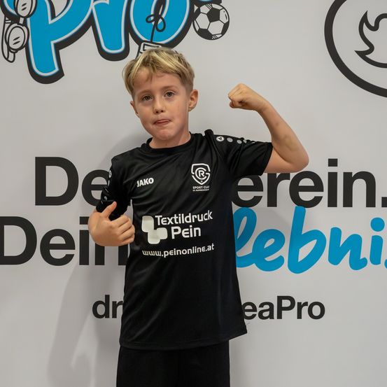 Ein junger Junge in einem schwarzen Fußballtrikot mit 'Textildruck Pein' und 'Jako' Logos steht vor einem weißen Hintergrund mit Fußballthema.