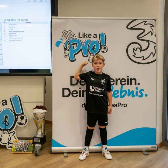 Ein junger Junge steht vor einem großen Banner mit einem Pokal und Büchern neben ihm, in einem schwarzen Fußballtrikot. Das Banner hat Text auf Deutsch und Englisch und enthält ein Logo.