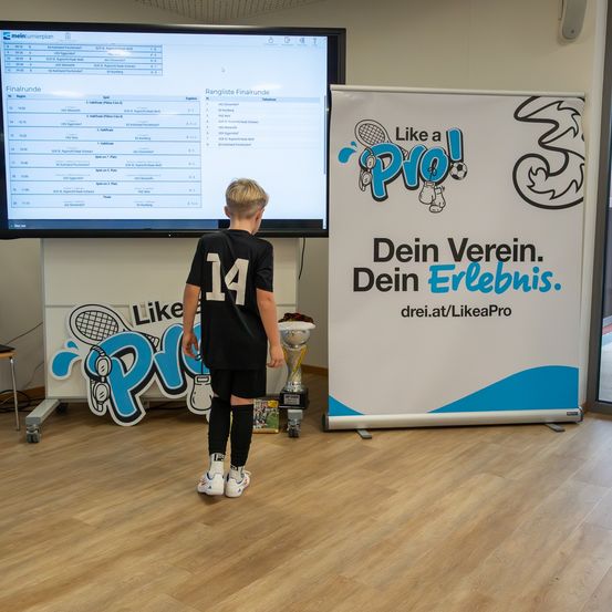Ein junger Junge in einem Fußballtrikot mit der Nummer 14 steht vor einem Monitor, der ein Sportereignis und einen Banner mit einem Sportthema zeigt.