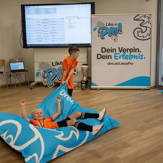 Zwei Kinder sind in einem Raum, eines liegt auf einem Sitzsack. Ein Junge steht hinter ihnen. Ein Monitor, Laptop und eine Trophäe befinden sich im Raum.