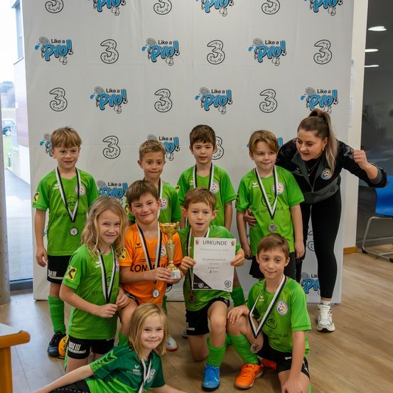 Eine Gruppe junger Fußballspieler in grünen Trikots und mit Medaillen posiert für ein Foto mit einer Trophäe und einem Zertifikat. Eine Frau steht hinter ihnen.