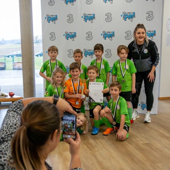 Eine Gruppe junger Fußballspieler posiert für ein Foto mit Medaillen und einem Zertifikat. Ein Kind hält einen Pokal. Eine Frau macht ein Foto von ihnen.