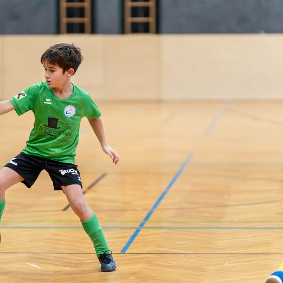 Ein junger Junge spielt Futsal auf einem Hallenplatz. Er trägt ein grünes Trikot mit Logo und dem Text Trijos Games. Er streckt sein Bein in Vorbereitung auf einen Schuss.
