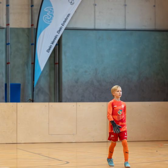 Ein junger Junge steht auf einem Hallenplatz und trägt ein orange-weißes Fußballtrikot. Er schaut nach links, wahrscheinlich wartet er auf den Beginn des Spiels. Hinter ihm befindet sich eine blaue Fahne mit Text.