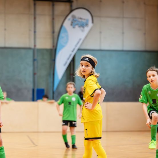 Eine Gruppe junger Kinder spielt Hallenfußball. Ein Kind in einem gelben Trikot schaut zur Seite. Hinter ihr sind zwei Kinder in grünen Trikots zu sehen. Ein Banner hängt an der Wand im Hintergrund.