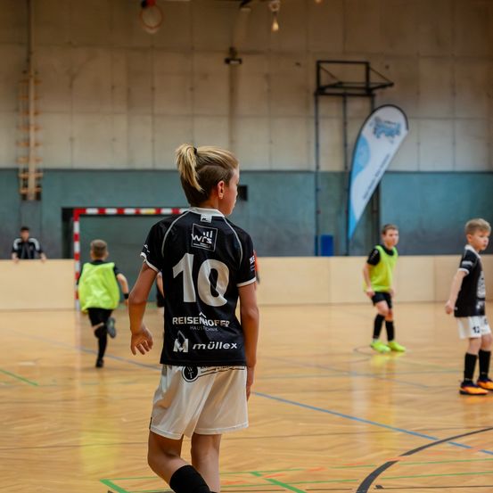 Eine Gruppe von Kindern spielt Fußball auf einem Hallenplatz. Ein Kind mit der Nummer 10 auf dem Trikot steht hinten.