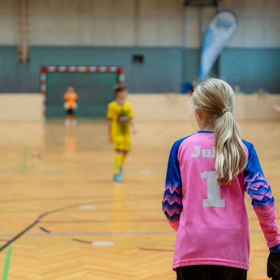 Ein junges Mädchen mit blonden Haaren, das ein rosa-blaues Trikot mit der Nummer 1 trägt, schaut sich ein Spiel auf einem Hallenplatz an. Im unscharfen Hintergrund ist ein Spieler in einem gelben Trikot zu sehen.