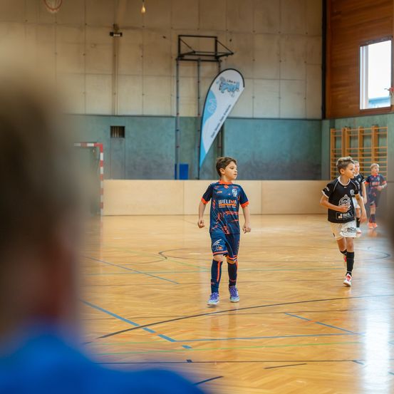 Zwei junge Jungen spielen Fußball auf einem Hallenplatz. Ein Junge in Blau schaut zurück, während ein anderer in Schwarz auf ihn zuläuft. Eine verschwommene Figur beobachtet von der Seite.