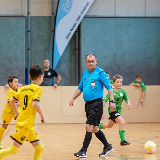 Ein Schiedsrichter läuft über das Spielfeld und beaufsichtigt Kinder in gelben und grünen Trikots. Ein Banner und mehrere Zuschauer sind im Hintergrund.