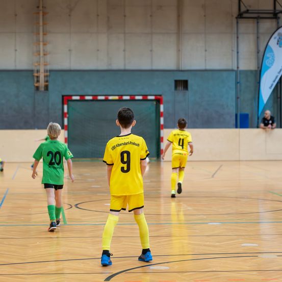Vier junge Spieler in gelben und grünen Trikots spielen Fußball auf einem Hallenplatz. Ein Spieler trägt die Nummer 9.