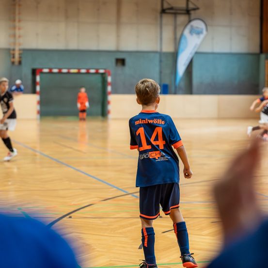 Ein junger Junge mit einem blauen Trikot mit der Nummer 14 steht auf einem Hallenbasketballplatz und schaut sich ein Spiel an. Andere Spieler sind im Hintergrund.