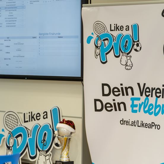 Ein Pokal auf einem Display-Board mit dem Slogan 'Like a Pro' und einem sportbezogenen Logo wird gezeigt. Dahinter zeigt ein Computerbildschirm eine Liste mit Namen und Punkten an.