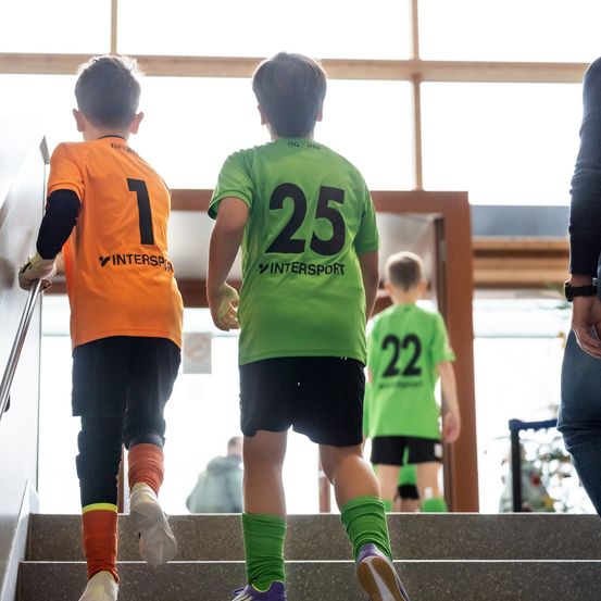 Drei junge Jungen in Fußballtrikots steigen eine Treppe hinauf, wobei der Junge in der Mitte ein grünes Trikot mit der Nummer 25 trägt.
