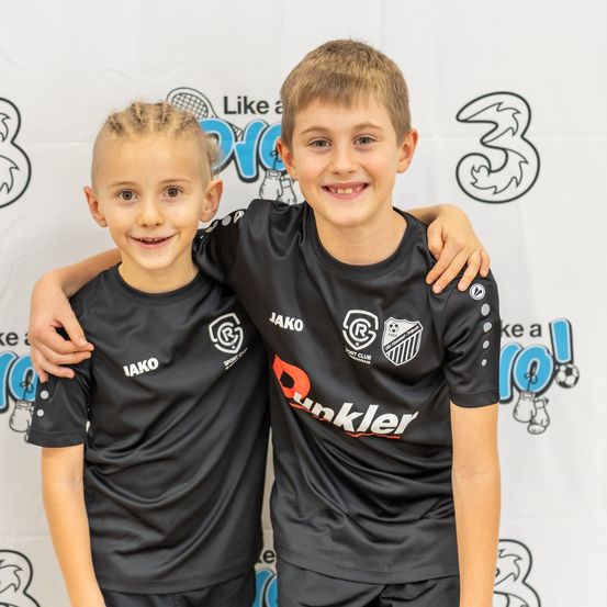 Zwei Jungen in schwarzen Fußballtrikots posieren für ein Foto, lächelnd und stehend nah beieinander. Der Junge auf der linken Seite hat geflochtene Haare, während der Junge auf der rechten Seite ein Logo auf seinem Trikot hat. Dahinter ist ein Hintergrund mit einer Nummer 3 und einem Logo.