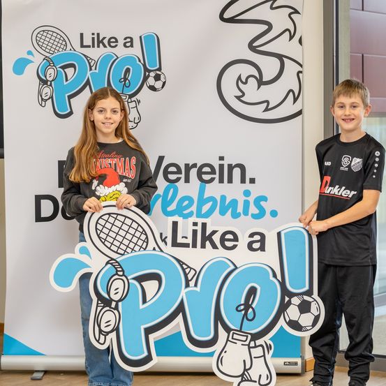 Zwei Kinder posieren neben einem Banner mit dem Slogan 'Wie ein Profi!' und Sportgeräten. Ein Mädchen hält ein Schild und ein Junge steht neben ihr.