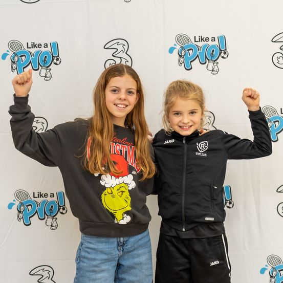 Zwei Mädchen stehen vor einem weißen Hintergrund mit einem Logo. Das Mädchen auf der linken Seite trägt ein Grinch-Sweatshirt, während das Mädchen auf der rechten Seite eine schwarze Jacke mit einem Logo auf der Brust trägt. Beide haben ihre Fäuste erhoben.