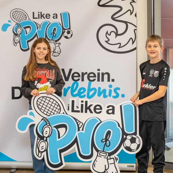 Zwei Kinder posieren neben einem Schild, das 'Like a Pro!' steht. Eines hält eine Tennisschläger, das andere einen Fußball. Sie stehen vor einem Banner mit Sportgrafiken.