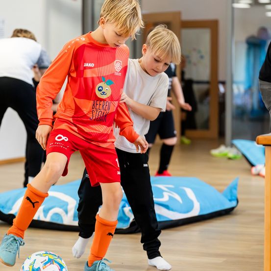 Zwei Jungen spielen Fußball in einer Turnhalle, einer in einem roten Trikot und der andere in einem weißen Hemd. Der Junge in Rot tritt den Ball.