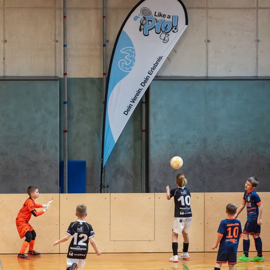 Hallensportplatz mit einem Banner mit der Aufschrift 'Wie ein Profi'. Mehrere junge Kinder spielen Fußball, ein Mädchen springt, um den Ball zu köpfeln.