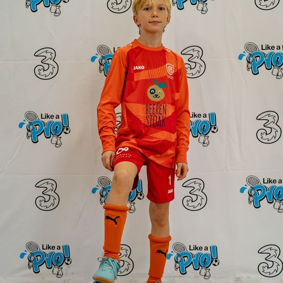Ein junger Junge in einem Fußballtrikot mit 'Jako' und 'BEEREN STOAK' darauf steht auf einer Bühne und lächelt. Er trägt orange Socken, blaue Turnschuhe und rote Shorts. Hinter ihm ist ein weißer Hintergrund mit '3' und 'Like a Pro!' Grafiken.